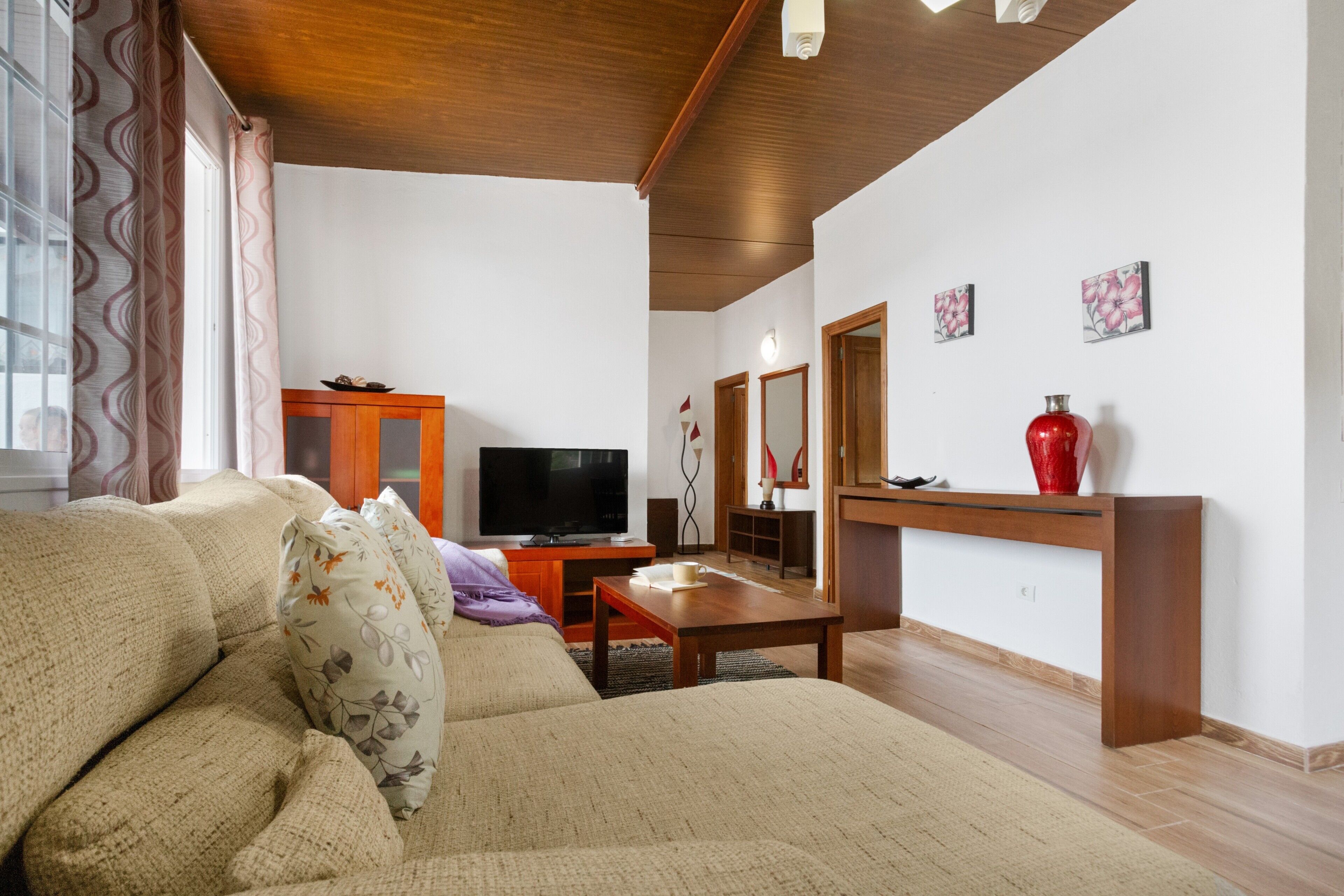 Een woonkamer met een bank, televisie, houten kast en een rode vaas op een console tafel.