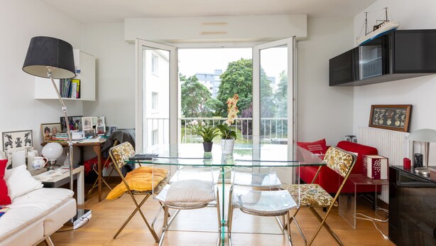 A modern living room with a glass dining table, a sofa, and a red chair. There is a bookshelf, a lamp, and a window with a view of trees and buildings. - La Ville Allis by Interhome (Saint Malo)
