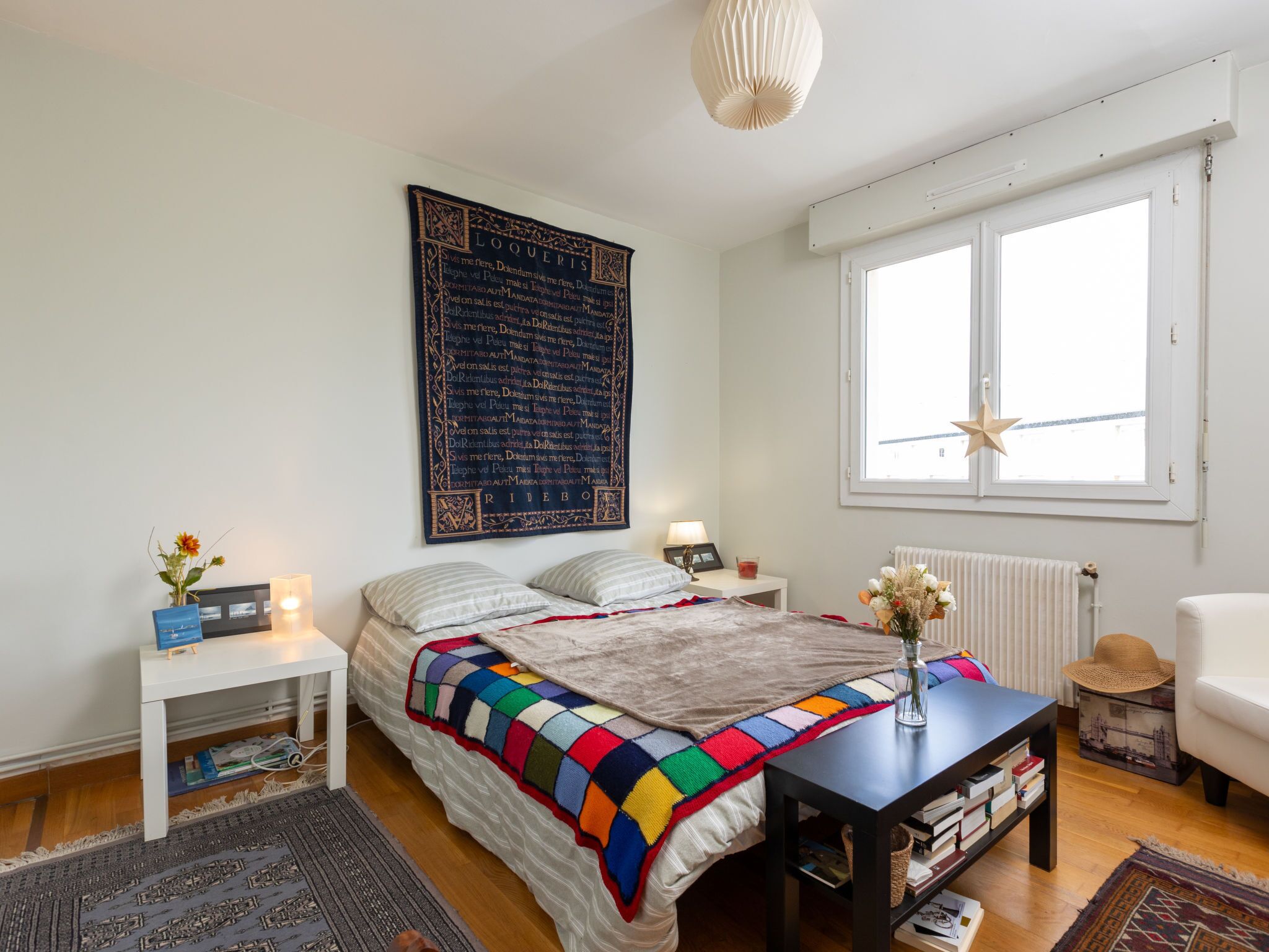 Une chambre avec un grand lit, une table de chevet avec une lampe, une fenêtre décorée d’une étoile et un radiateur.