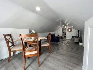 Ein Essbereich mit einem Glastisch und Holzstühlen, einer Deckenleuchte und Blick in ein Schlafzimmer mit Bett und Fernseher.