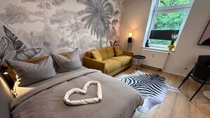 Quarto com cama, sofá, tapete de zebra, painel na parede, luminária de chão e janela com vista para vegetação.