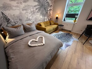 A bedroom with a bed, a sofa, a zebra rug, a wall mural, a floor lamp, and a window with a view of greenery.