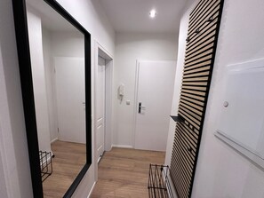A narrow hallway with white walls, wooden flooring, and a large mirror on the left. There are two doors, one with a light switch and another with a door handle.