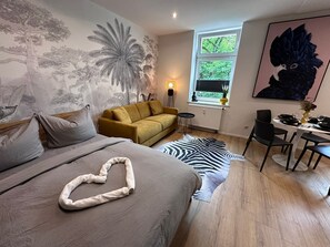 A bedroom with a bed, a sofa, a zebra rug, a desk with chairs, and a window with a view of trees.