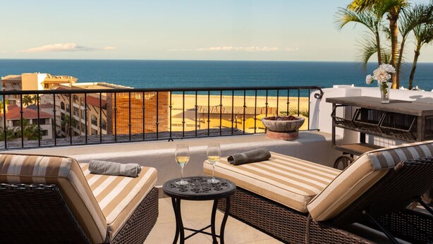 A balcony with wicker furniture, a small table with two wine glasses, and a view of the beach and ocean.