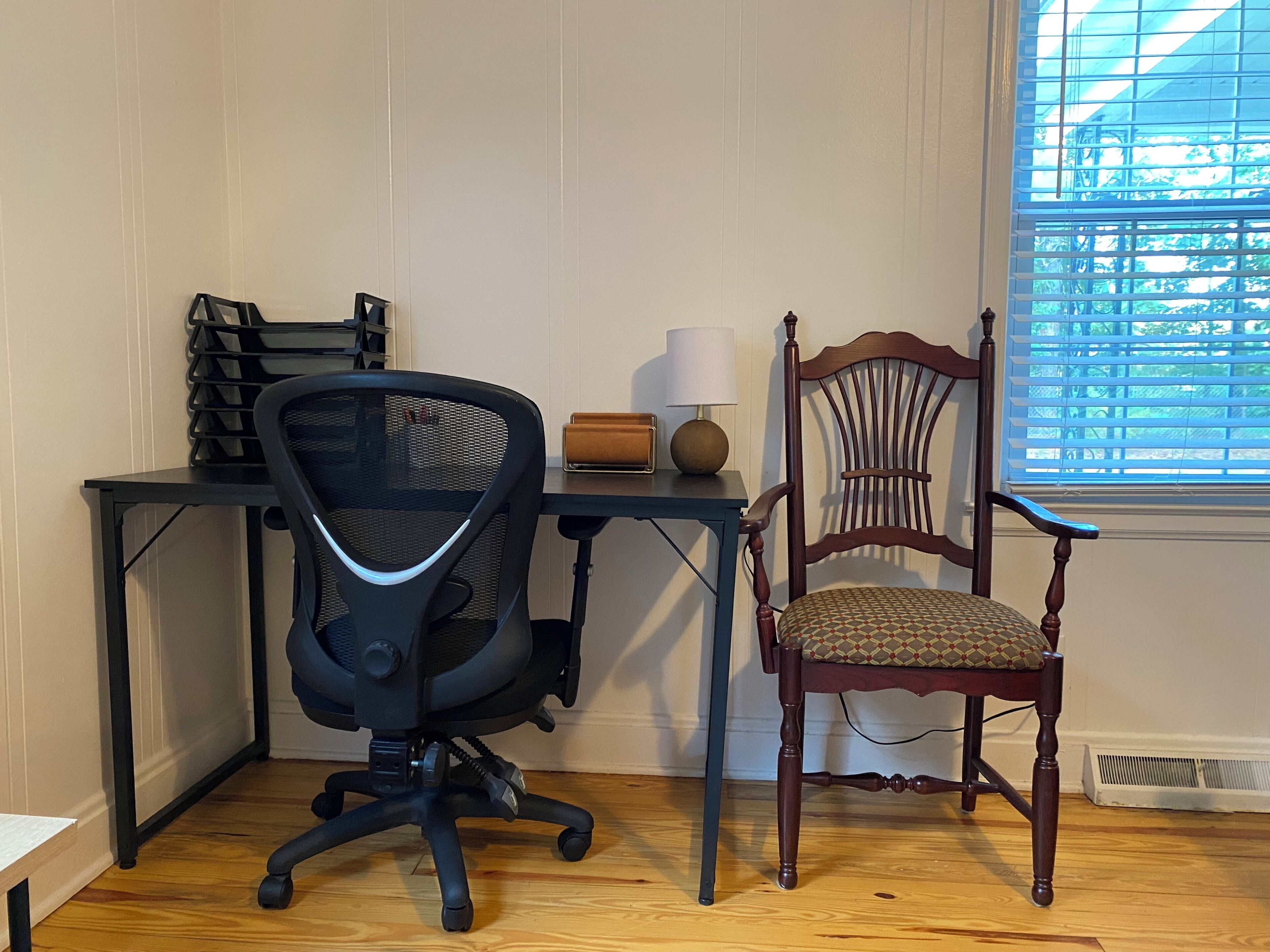 A room with a wooden desk, a black office chair, a wooden chair with a patterned cushion, and a window with blinds.