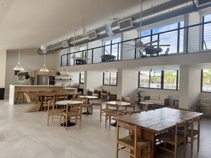 Un vaste espace de salle à manger avec plusieurs tables et chaises, un coin cuisine équipé d’appareils modernes et de grandes fenêtres laissant entrer la lumière naturelle.