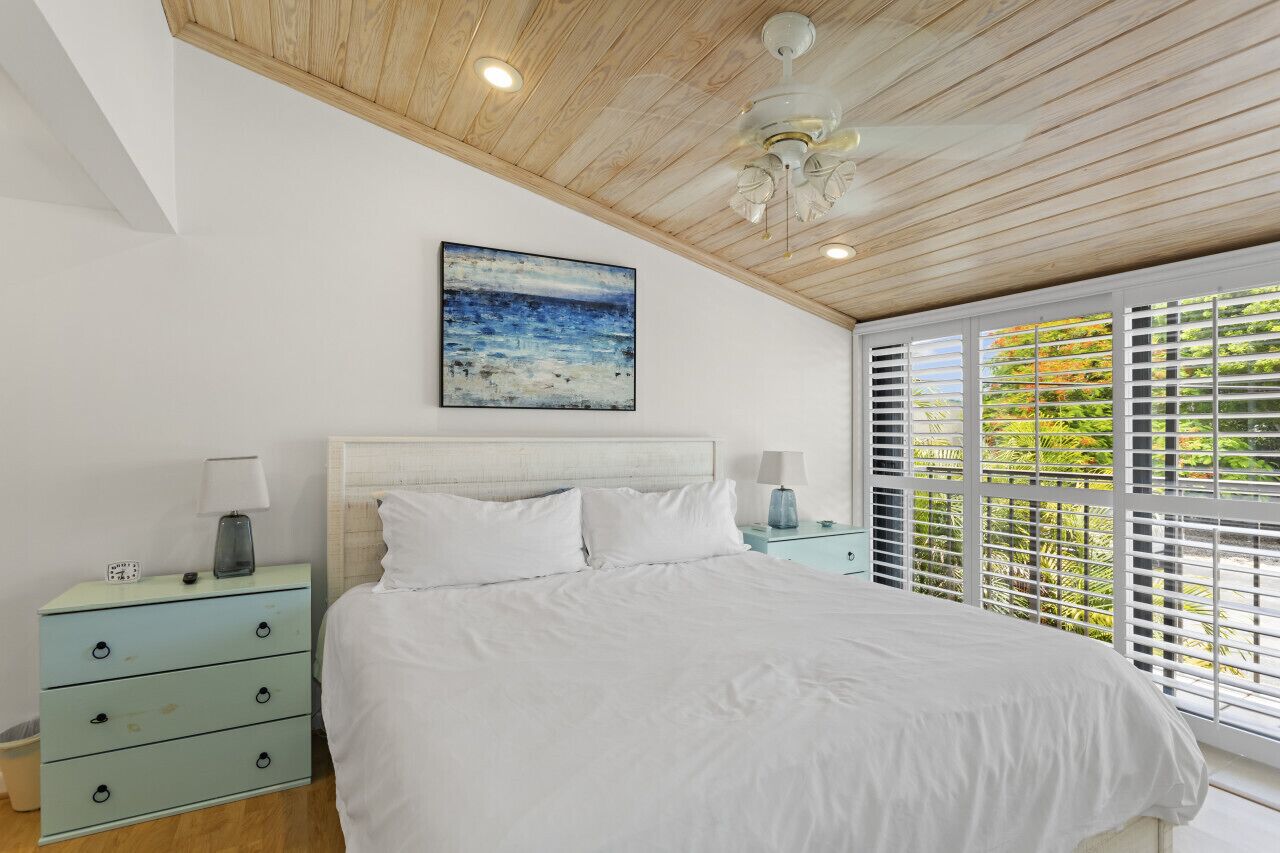 A bedroom with a wooden ceiling, a large bed with white bedding, a nightstand with a lamp, and a window with blinds.