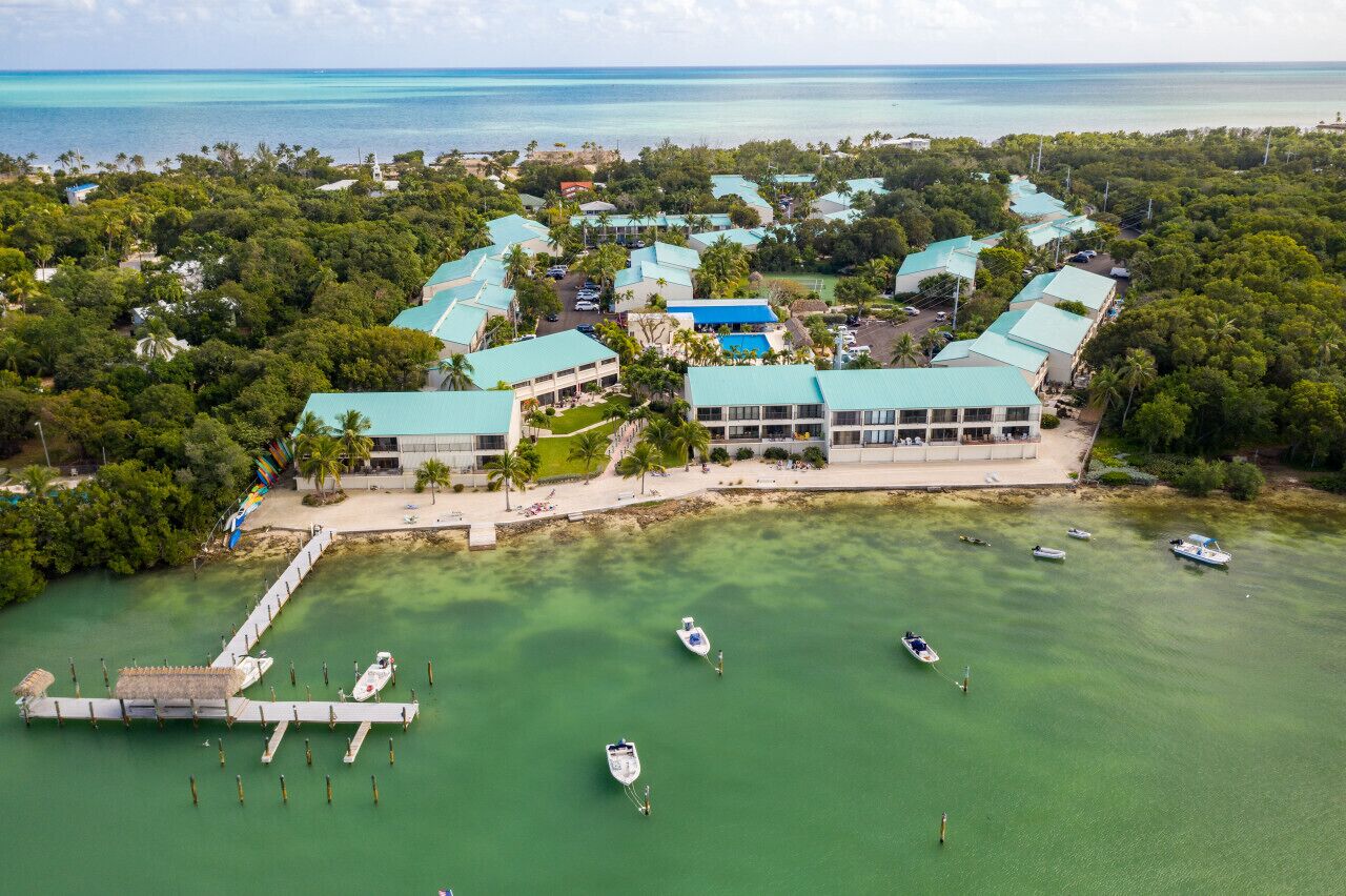 An aerial view of a resort with multiple buildings, a dock with boats, and a clear view of the ocean.