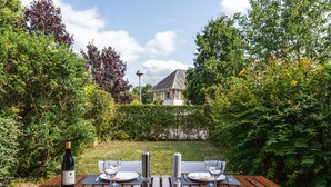 An outdoor dining table set with plates, glasses, cutlery, and a bottle of wine, surrounded by lush greenery and a house in the background. - La Pommeraie by Interhome (Villers sur mer)