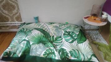 Una habitación con una cama cubierta por un edredón estampado con motivos de hojas tropicales, una mesita con un vaso y una figurita, y una pared decorada con un diseño.