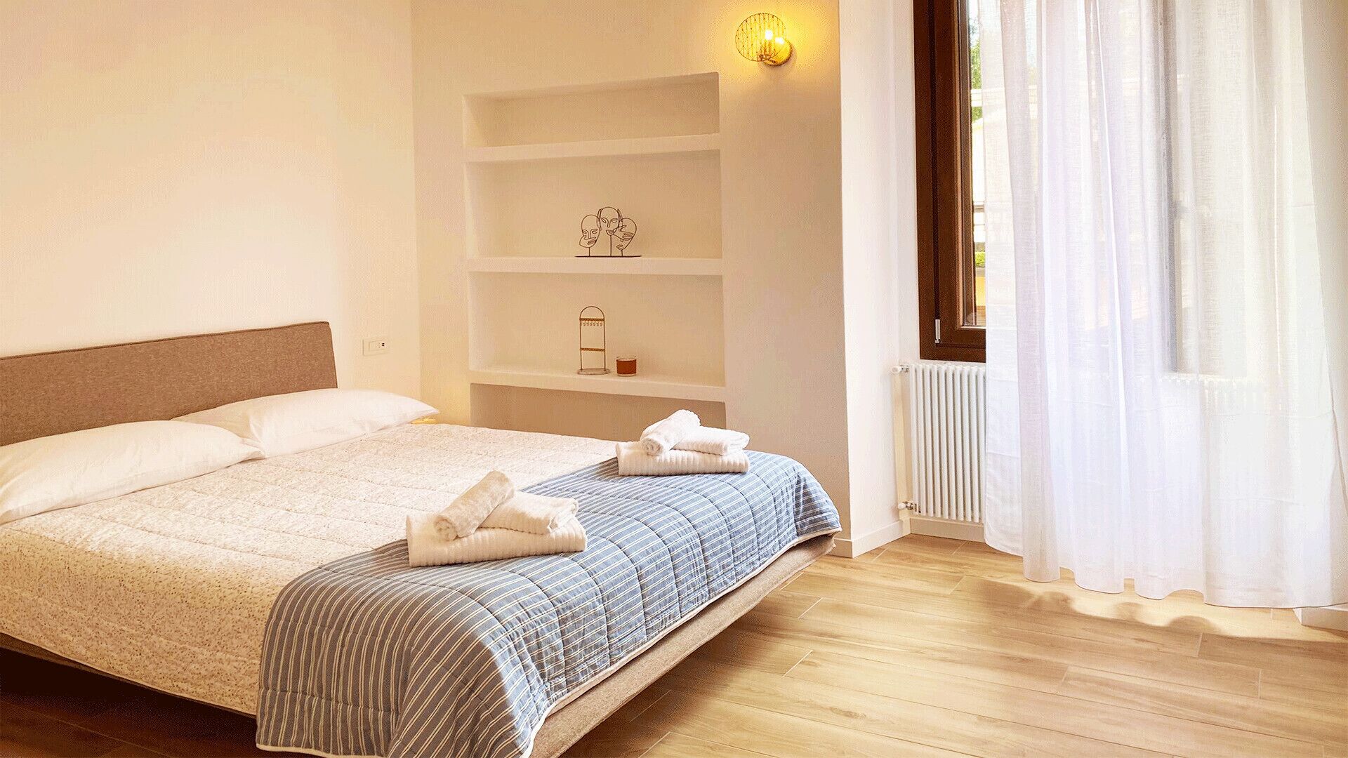 Un lit bien fait, avec une couverture à rayures, une tête de lit et une étagère ornée d’un objet décoratif et d’une bougie.
