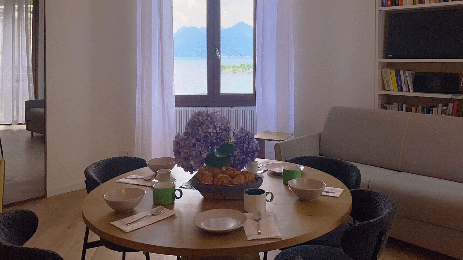 Zona pranzo con un tavolo imbandito per due persone, un vaso di fiori e vista sulle montagne attraverso una finestra.