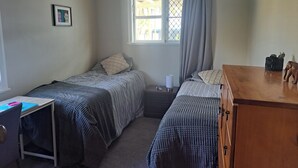 A bedroom with two single beds, a desk, a dresser, a window with curtains, and a small table with a lamp.