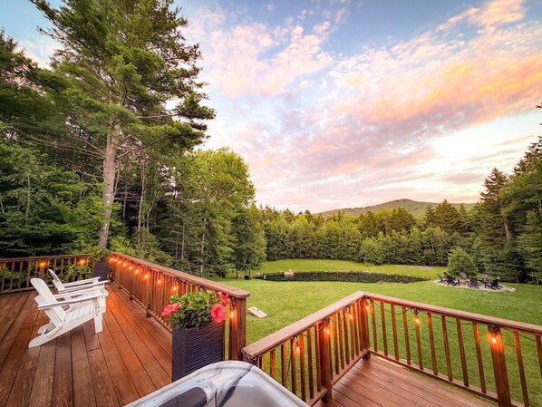 Une terrasse en bois dotée d’une balustrade, qui donne sur une cour verdoyante avec des arbres et une colline en arrière-plan.