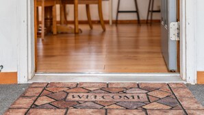 Un paillasson sur lequel est inscrit le mot « BIENVENUE », placé dans un vestibule carrelé qui mène à une pièce au plancher de bois et où se trouve une chaise.