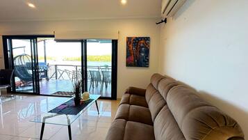 A spacious living room with a large beige sofa, a glass coffee table, and a view of a balcony with outdoor furniture.
