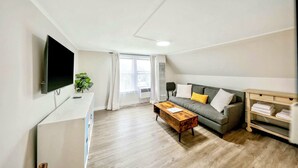 A modern living room with a flat-screen TV, a grey sofa with cushions, a wooden coffee table, and a wooden shelf with folded towels. - Chic, Cozy & New! Great location, Street Parking with AC (Renton)