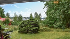 A view of a garden with a large coniferous tree, a red building, and a body of water in the background.