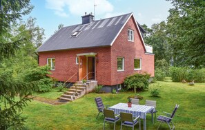 A red brick house with a sloped roof, a front door, and a small porch. There are steps leading to the door and a table with chairs outside.