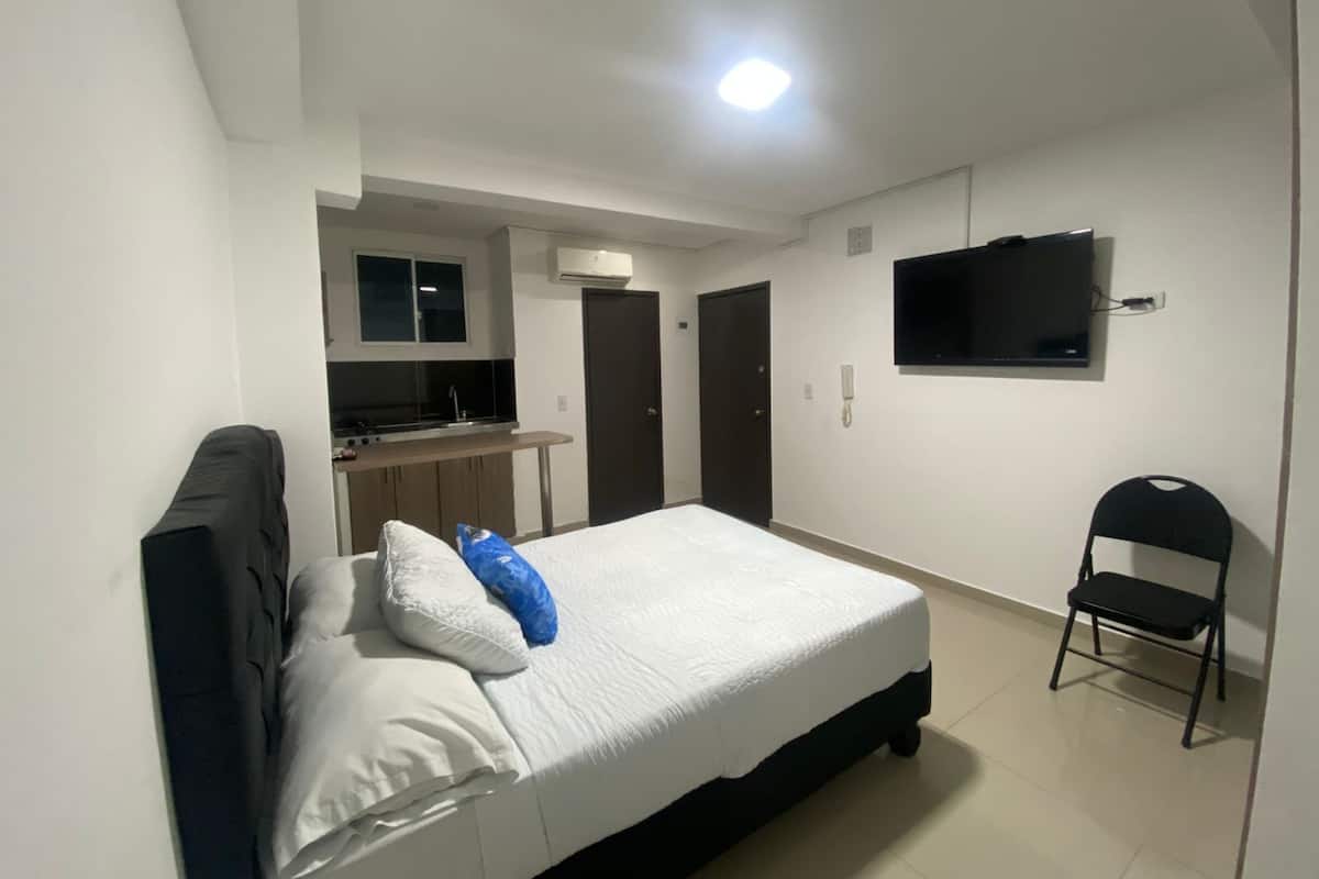 Een slaapkamer met een bed, een stoel, een televisie en een kitchenette op de achtergrond.