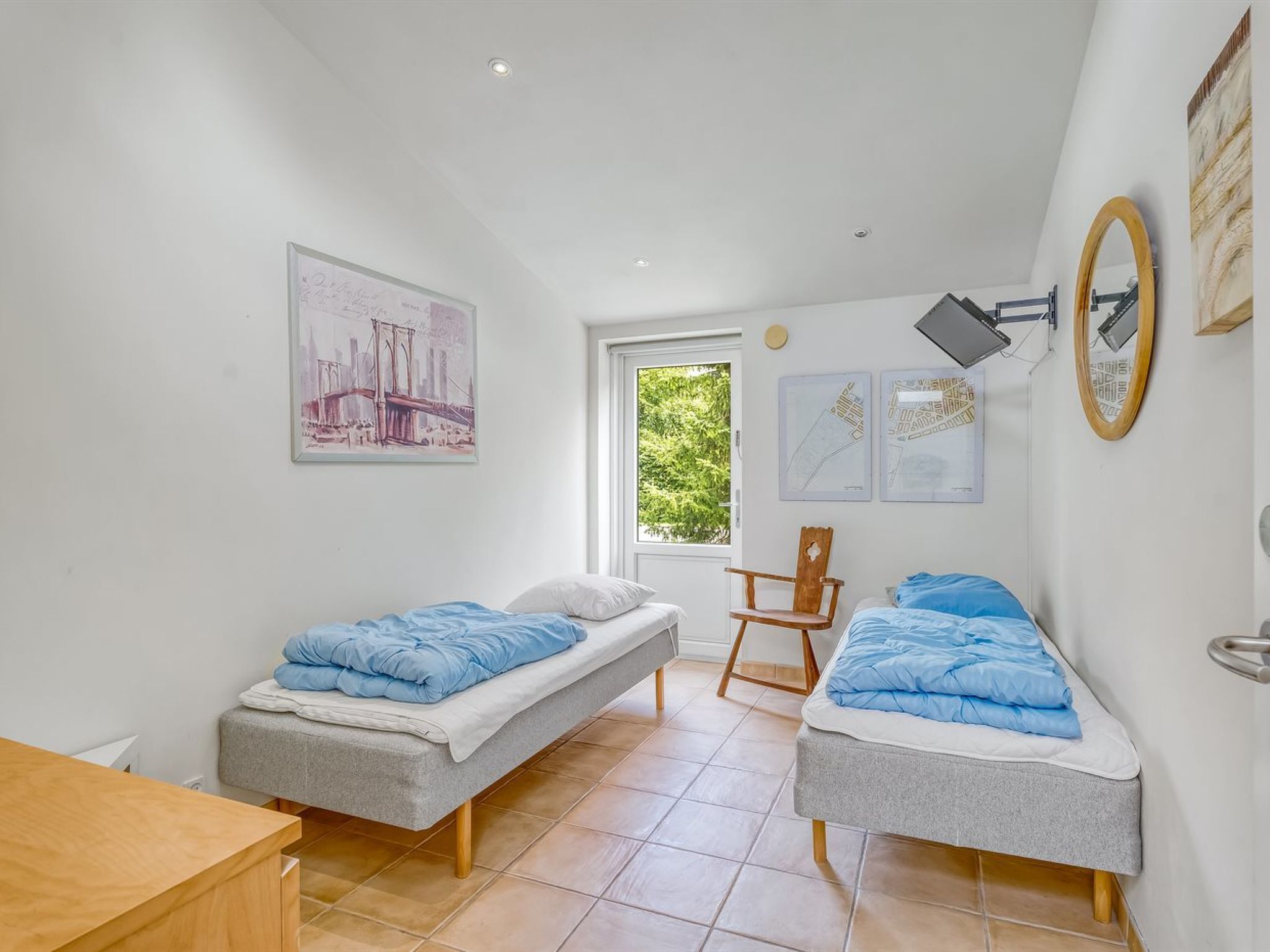 A small, bright room with two single beds, a wooden chair, and a desk.