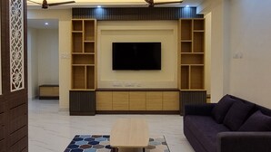 A modern living room with a flat-screen TV, built-in shelves, a ceiling fan, and a patterned rug. - Airport Inn  (Chennai)