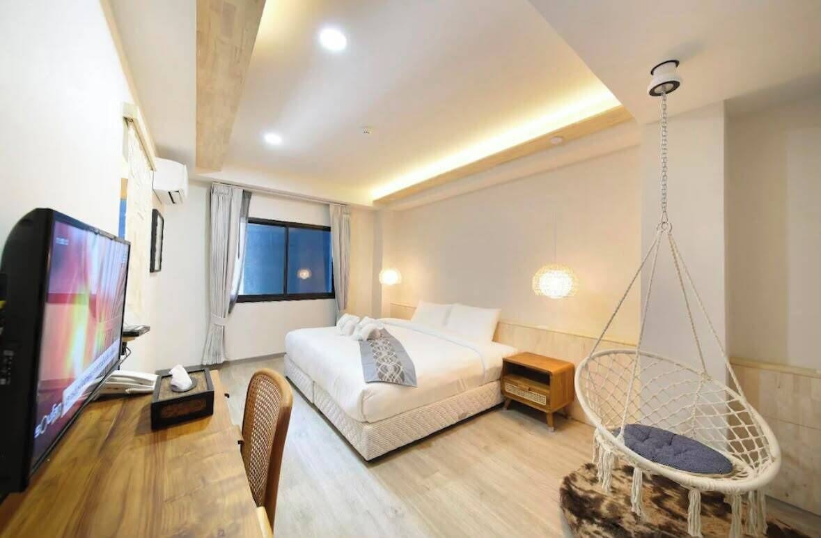 A modern hotel room with a bed, a desk with a computer, a hanging chair, and a window with curtains.