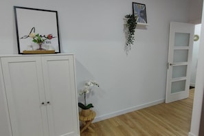 Une pièce avec une armoire blanche, un plancher en bois, une porte blanche et une étagère murale ornée d’un tableau encadré et d’une plante suspendue.