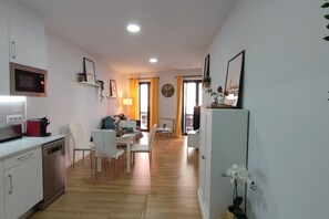 A modern kitchen with a microwave, dishwasher, and a coffee machine. There is a dining area with a table and chairs, and a living area with a sofa and a TV.