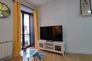 A living room with a flat-screen TV on a white stand, a clock on the wall, and a window with yellow curtains.