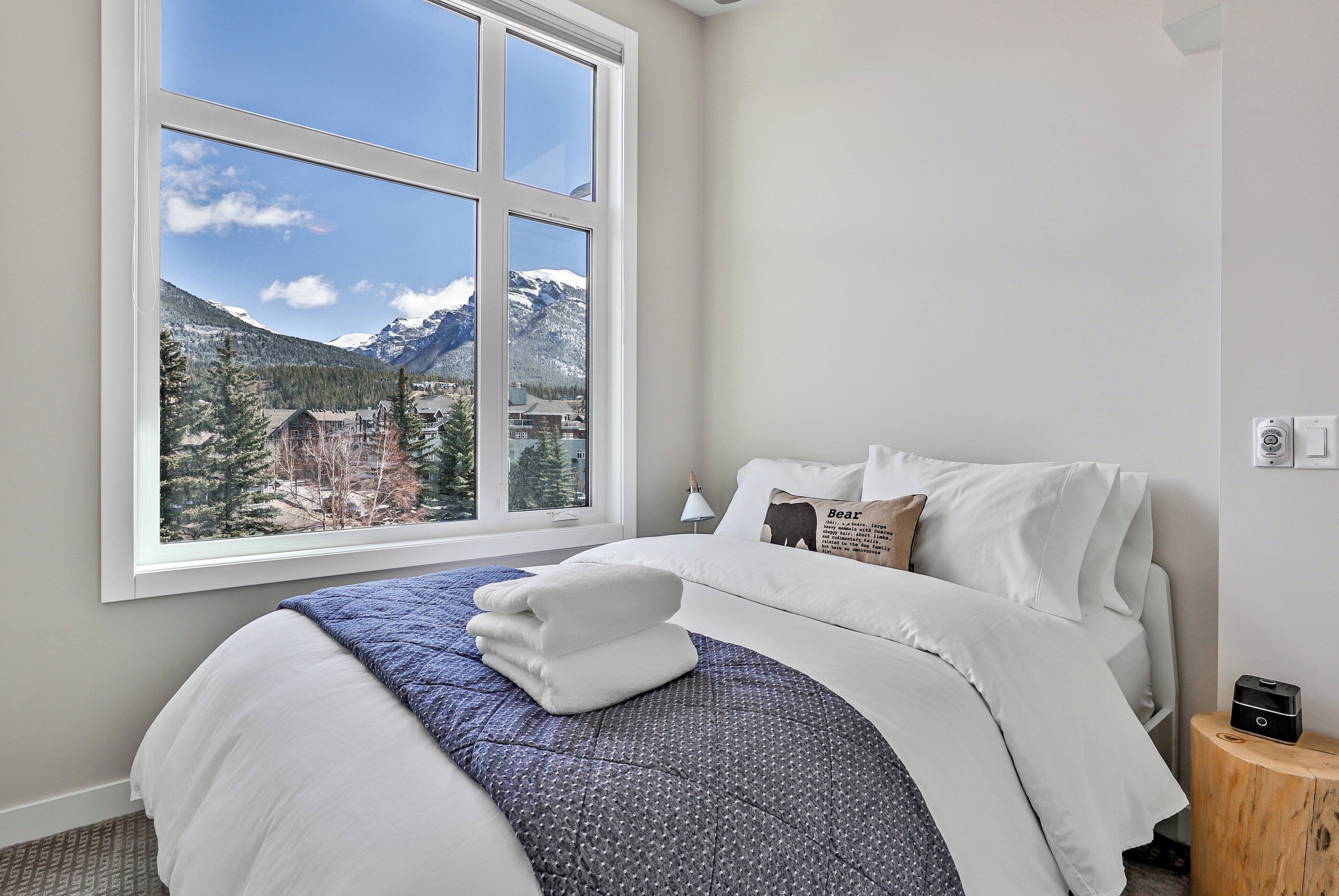 Une chambre d’hôtel dotée d’une grande fenêtre donnant sur les montagnes, d’un lit soigneusement fait avec du linge de maison blanc, et d’une petite table de chevet avec une lampe.
