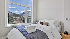 A hotel room with a large window offering a mountain view, a neatly made bed with white linens, and a small bedside table with a lamp.