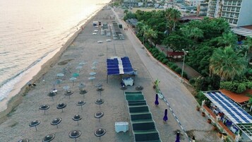 Vista aérea de una playa con filas de sombrillas y sillas de descanso, un área de estacionamiento y un edificio de techo plano al fondo.