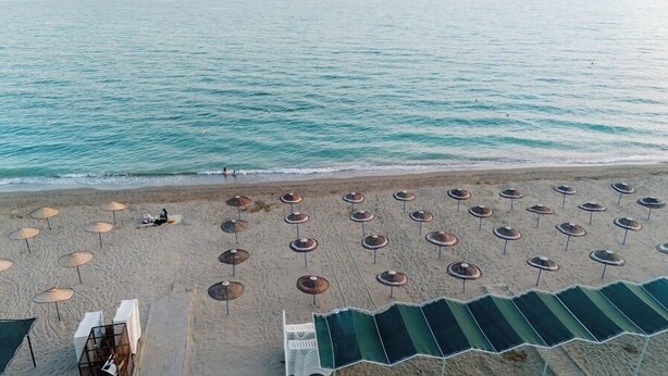 Vue aérienne d’une plage avec des rangées de parasols et de chaises de plage, un rivage sablonneux et une mer calme.
