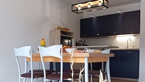 Uma área de jantar moderna com uma mesa de madeira e cadeiras brancas, um vaso azul com plantas secas e uma cozinha ao fundo.