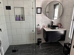Un baño moderno con cabina de ducha de vidrio, un tocador negro con espejo redondo y suelo de baldosas de mármol.