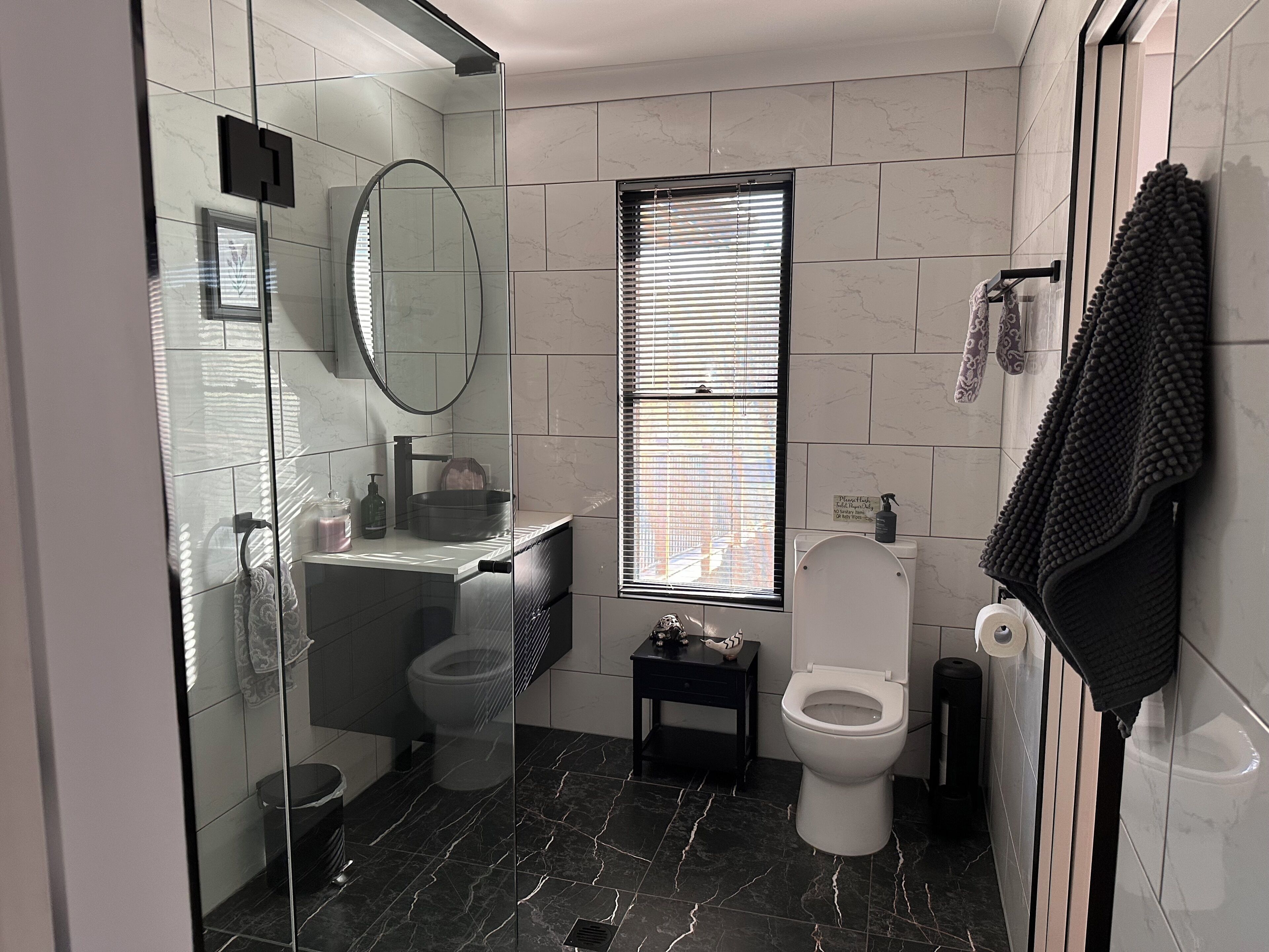 Un bany amb una cabina de dutxa de vidre, un vàter, un lavabo amb mirall i una finestra amb persianes.