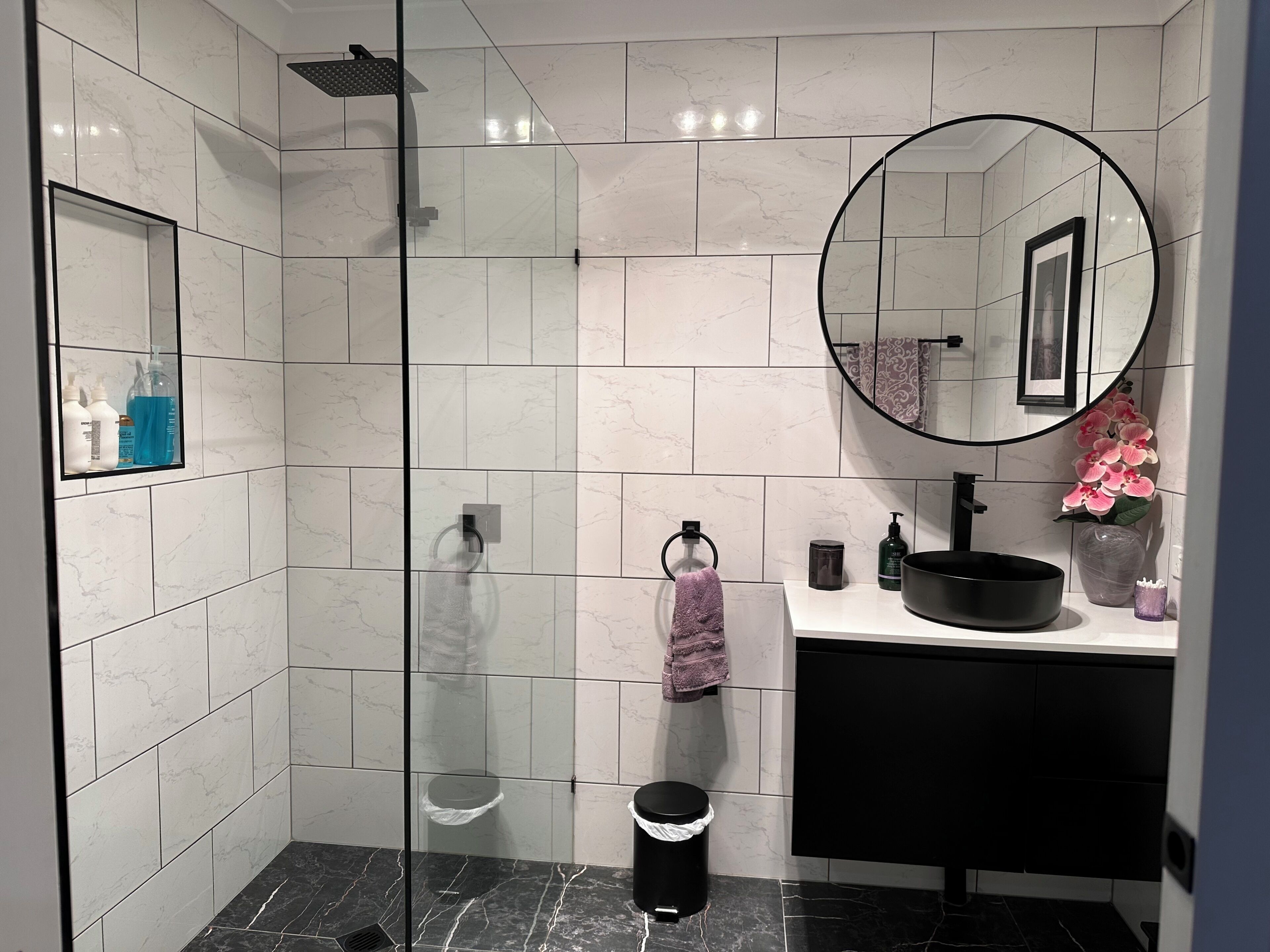 A modern bathroom with a glass shower enclosure, a round mirror, a black sink, and a marble-tiled floor.