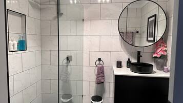 A modern bathroom with a glass shower enclosure, a round mirror, a black sink, and a marble-tiled floor.