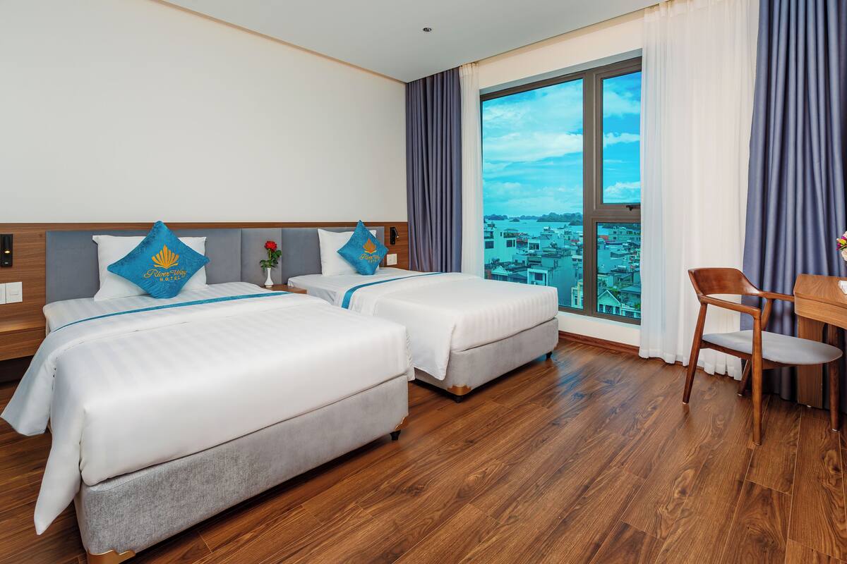 A hotel room with two beds, a wooden floor, a large window with a city view, and a desk with a chair.