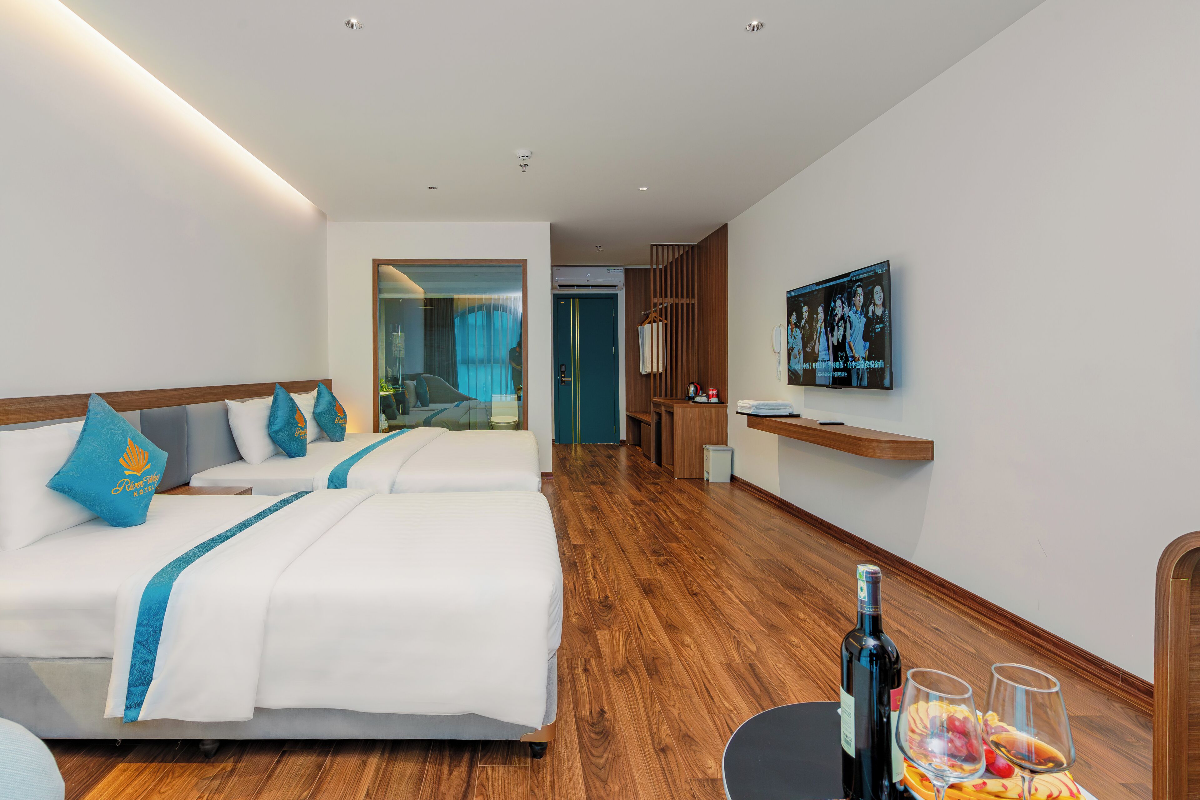 Habitación de hotel con dos camas, un televisor, una mesita con una botella y vasos, y piso de madera.