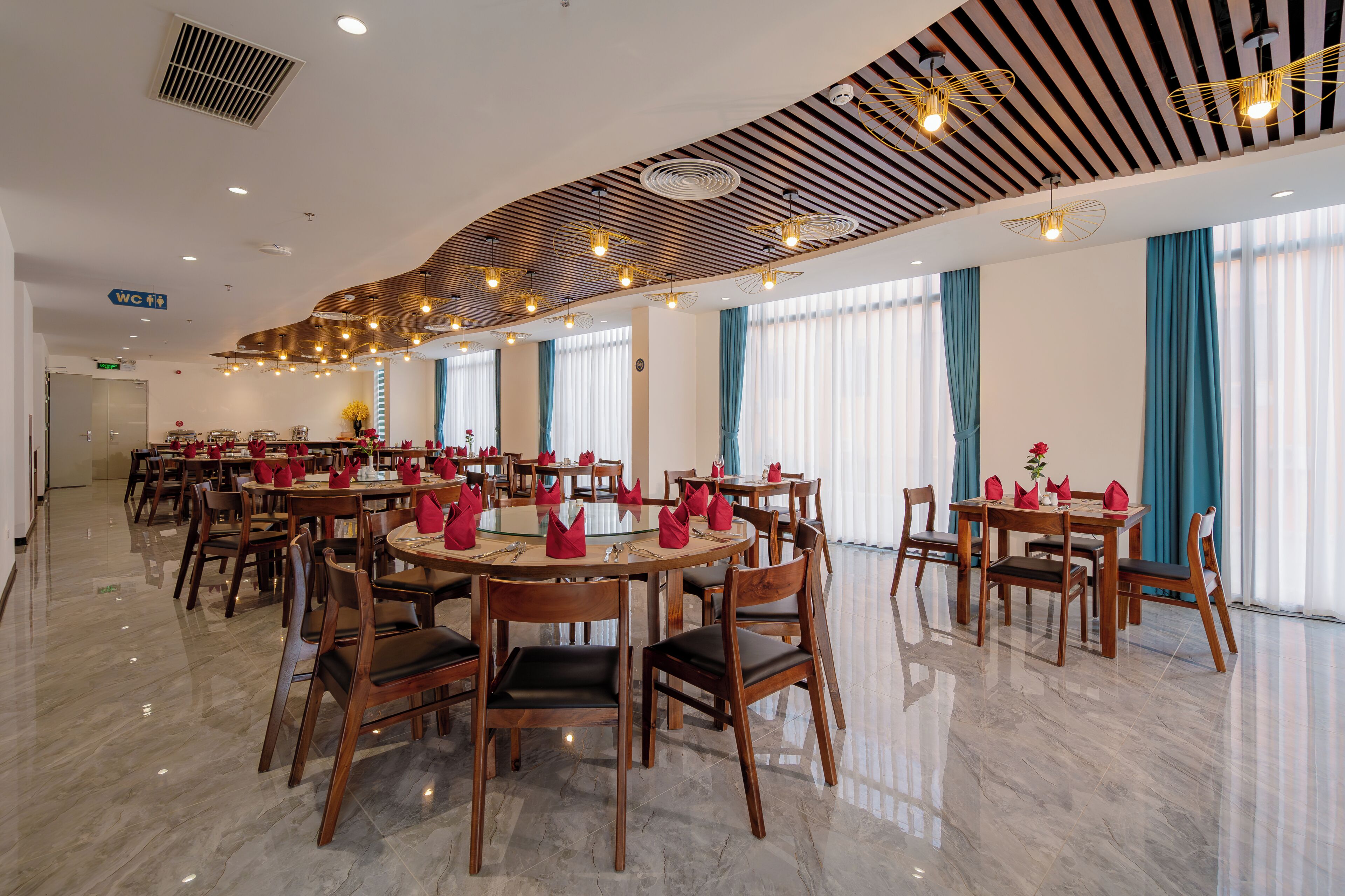 A hotel dining area with round tables, wooden chairs, and modern ceiling lighting.