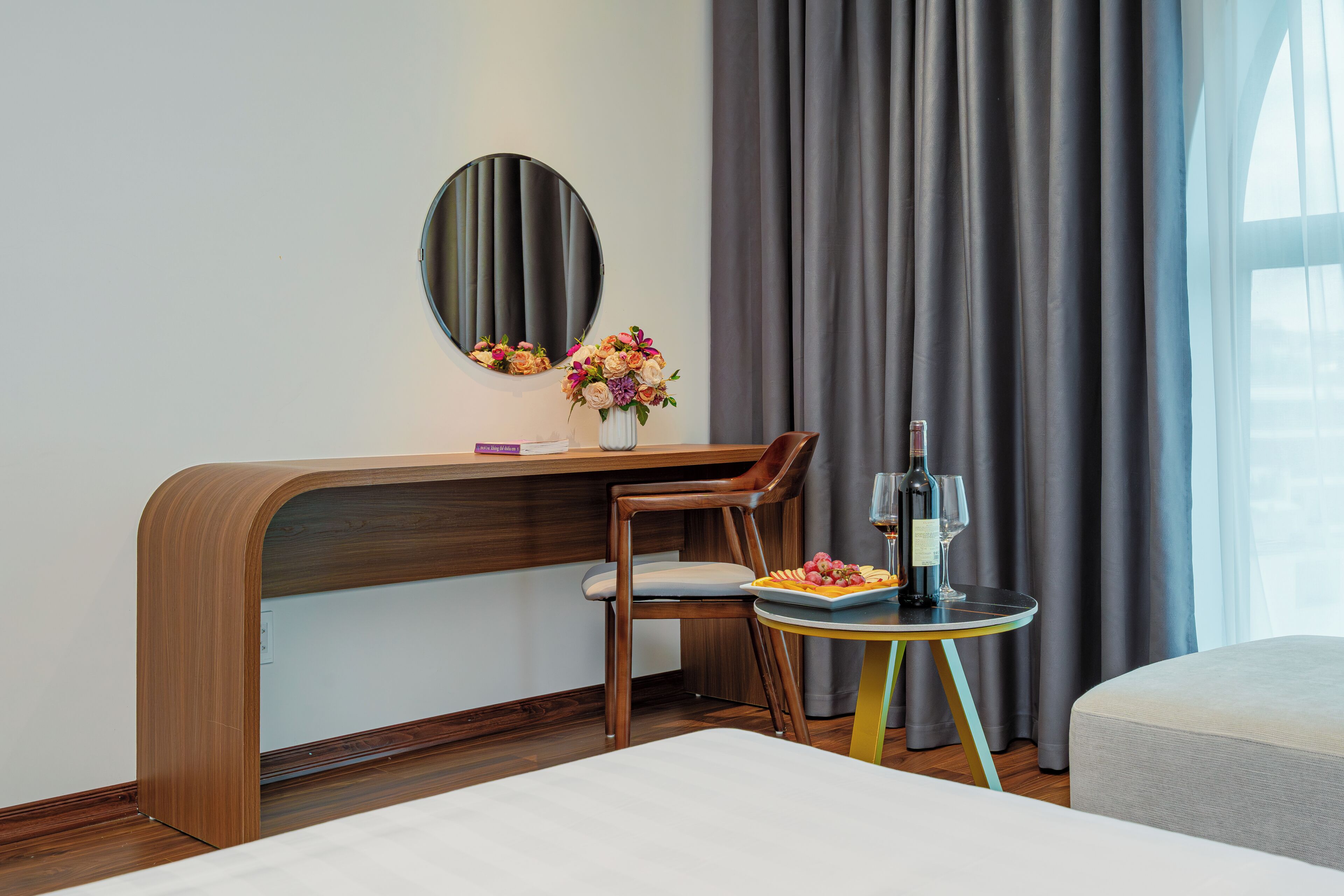 Habitación de hotel con un escritorio de madera, una silla, una mesita redonda con una botella y vasos, y una cama.