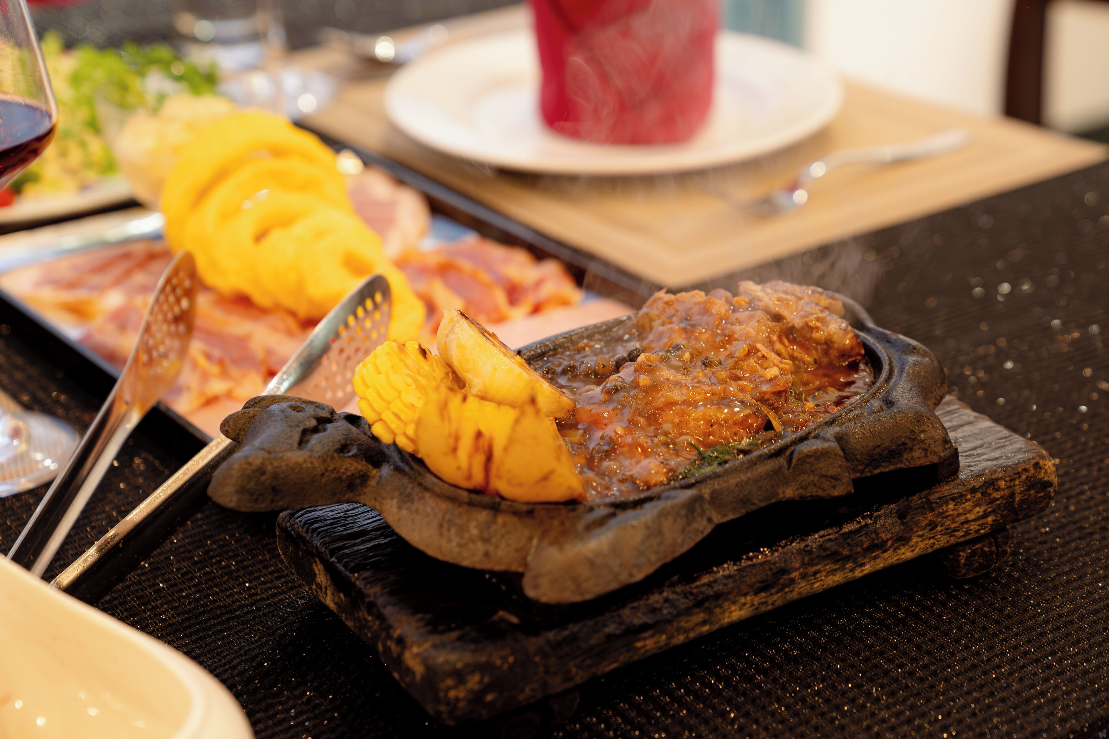 A dining table with a hot plate containing food, a glass of red drink, and a plate with sliced yellow fruit.