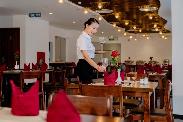 A restaurant with wooden tables and chairs, red napkins, and a waiter serving a glass of wine. - River Way Hotel Ha Long (Ha Long)