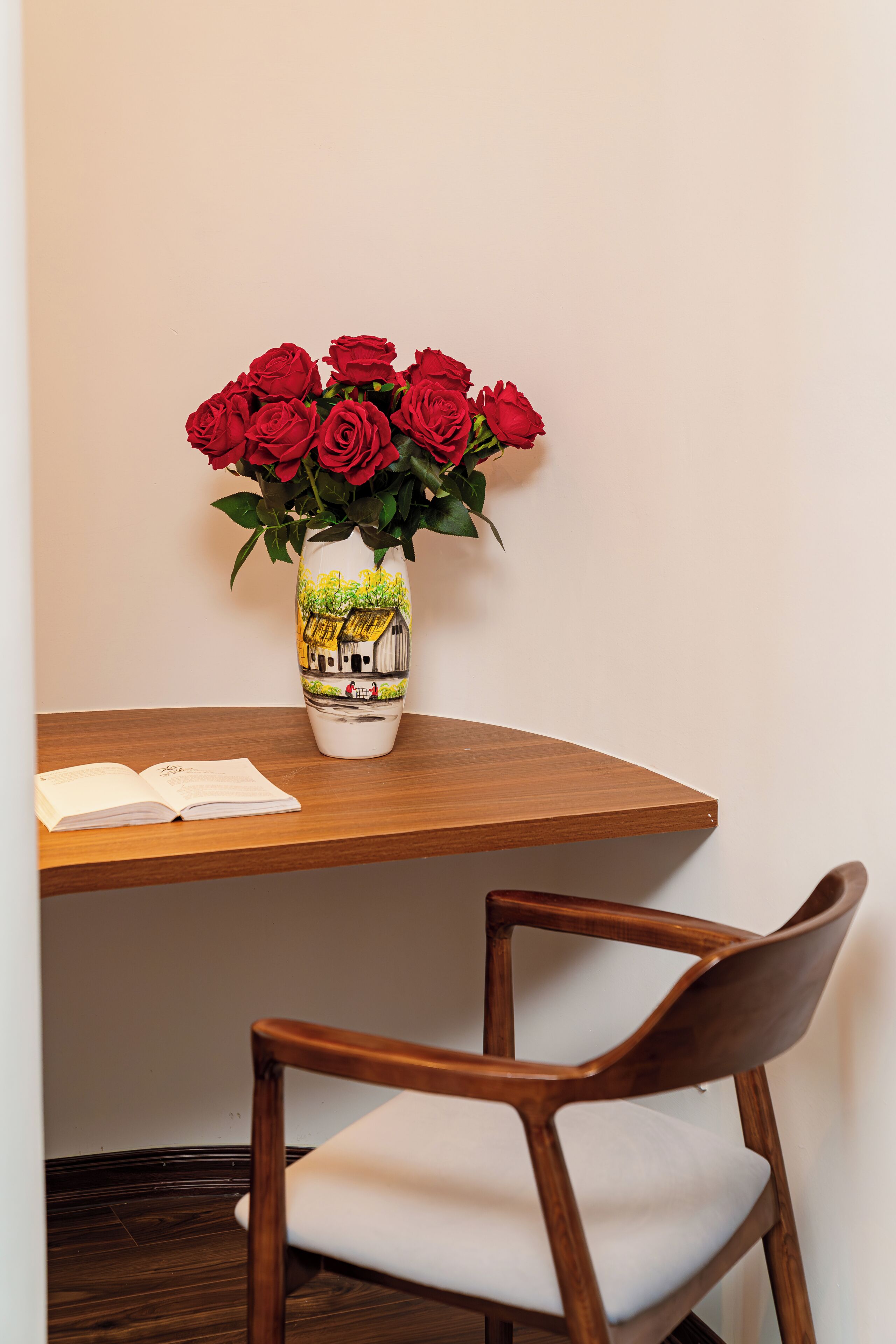 A wooden table with a white chair, a vase with red roses, and a book on the table.