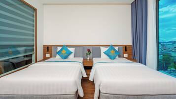 Habitación de hotel con dos camas, una mesita de noche, un florero con una flor y un ventanal con vistas a la ciudad.