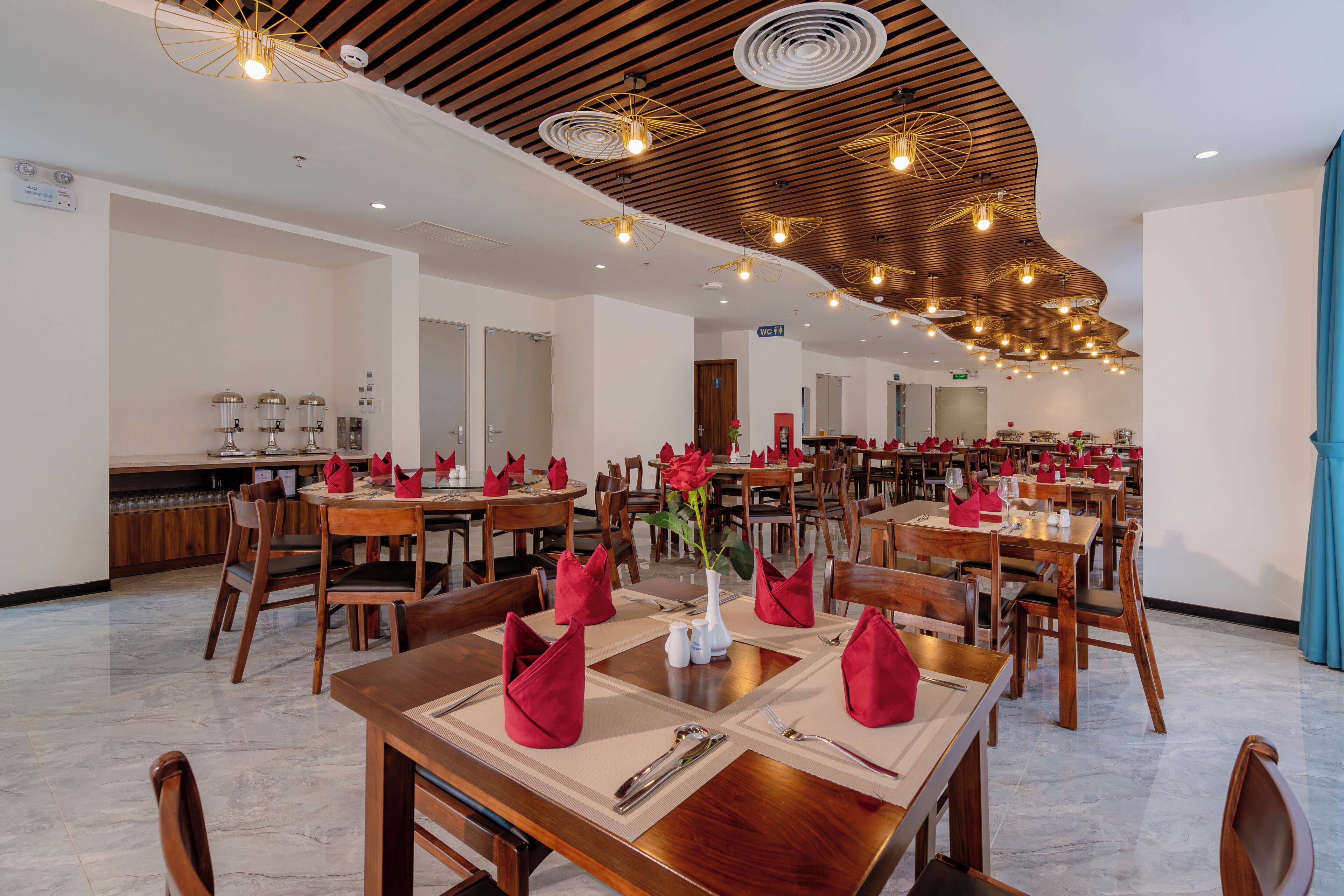 A spacious dining area with wooden tables, red napkins, and modern pendant lighting.