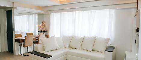 A modern living room with a sectional sofa, a dining table with chairs, and a large window with curtains.